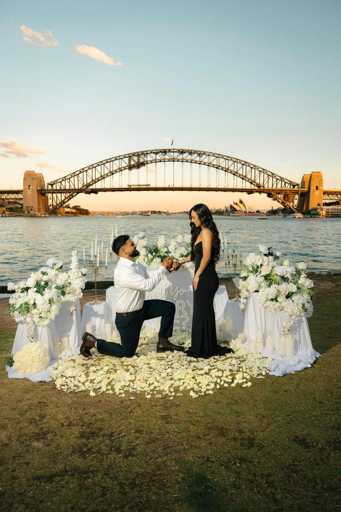 Jaz on one knee before Rianna among white flowers, the Harbour Bridge behind them